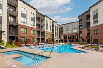 A large swimming pool is surrounded by lounge chairs in front of apartment buildings.